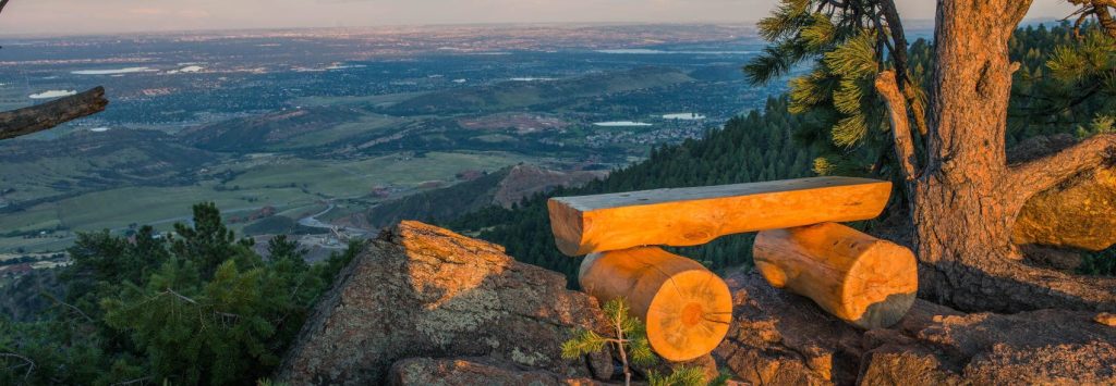 Relaxing Colorado mountain view from a wooded site, showcasing a sturdy wooden bench on rocky terrain with lush trees, capturing tranquility and natural beauty perfect for scenic real estate marketing.