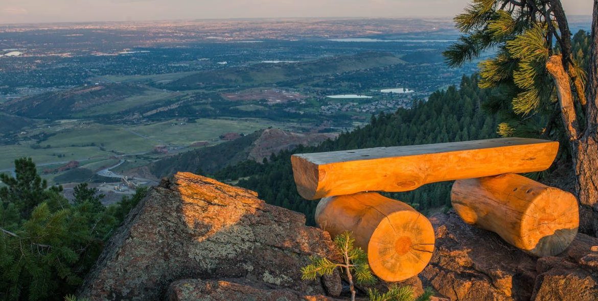 Relaxing Colorado mountain view from a wooded site, showcasing a sturdy wooden bench on rocky terrain with lush trees, capturing tranquility and natural beauty perfect for scenic real estate marketing.