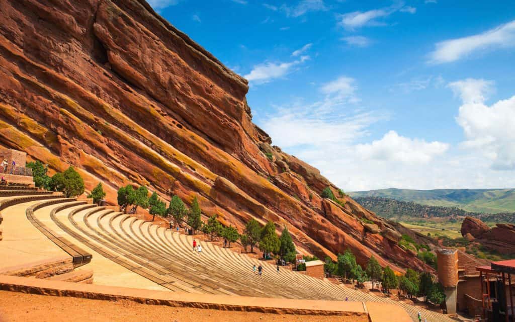 Vibrant daytime scene at Red Rocks Amphitheatre showcasing iconic red sandstone formations, rows of seating, visitors exploring the area, and clear blue sky, highlighting its status as a premier outdoor concert destination.